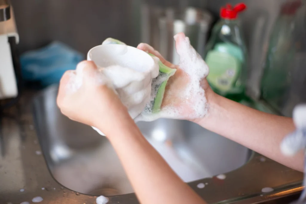 Hands washing a white dish with a sponge and soap suds over a kitchen sink